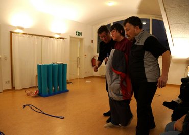 Zwei Männer links und rechts stützen eine Dame in der Mitte, die einen Schlafsack hält und eine Glasflasche.