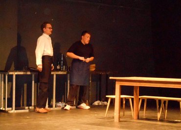 Zwei Darsteller in Schürzen stehen vor einem Tisch.