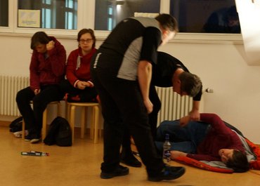 Eine Person liegt in einem Schlafsack, zwei weitere Personen gehen auf sie zu, einer nimmt ihren Arm.