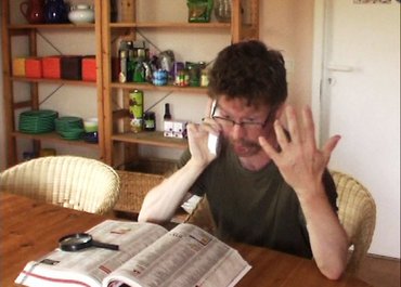 Mann telefoniert und hält eine Hand erhoben, vor ihm liegt ein Katalog mit einer Lupe.