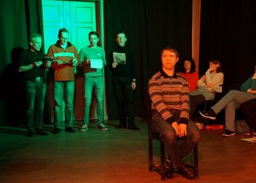 Ein Mann sitzt auf einem Stuhl in der Mitte des Raumes, im Hintergrund stehen und sitzen mehrere Personen.