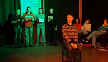 Ein Mann sitzt auf einem Stuhl in der Mitte des Raumes, im Hintergrund stehen und sitzen mehrere Personen.