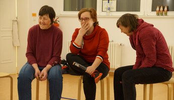 Drei Damen lachen, während sie nebeneinander auf Hockern sitzen.