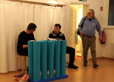 Zwei Personen sitzen auf Stühlen und sprechen miteinander, während eine weitere Person den Raum betritt.