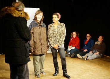 Darsteller sitzen im Hintergrund, während im Vordergrund drei Darsteller ein Gespräch führen.