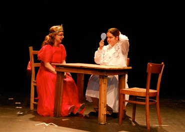 Zwei Damen sitzen an einem Tisch, eine trägt eine Krone, die andere blickt in einen Handspiegel.