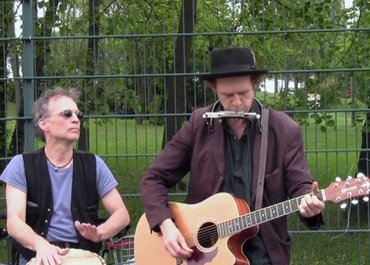 Zwei Männer stehen draußen vor einem Zaun, einer trommelt, der andere spielt Gitarre.