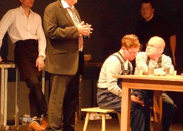 Mann im Anzug spricht und blickt Richtung Publikum, während Personen im Hintergrund stehen oder sitzen.