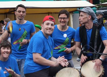 Fünf Personen aus der Trommelgruppe spielen gemeinsam, eine Person lächelt in die Kamera.