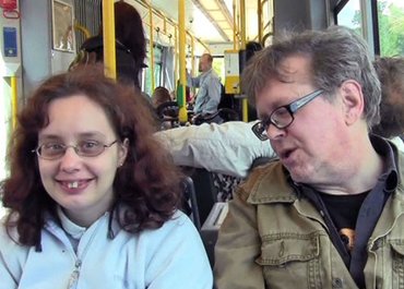 Frau und Mann sitzen nebeneinander in einer Straßenbahn, Frau lächelt in die Kamera, Mann spricht mit ihr.