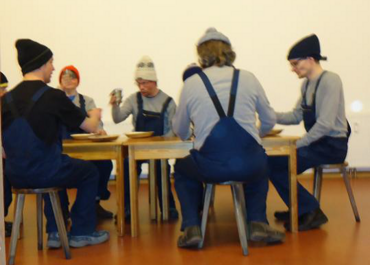 Personengruppe in Latzhosen sitzt an einem Tisch und stößt mit Getränken an.