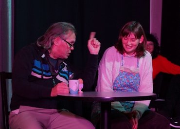 Zwei Personen sitzen nebeneinander an einem runden Tisch, ein Mann zeigt mit dem Finger und spricht