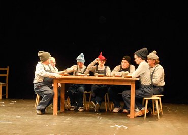 Sieben Darsteller mit Mützen und Latzhosen sitzen an einem Esstisch mit Tellern und Bechern.