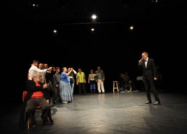 Rechts singt ein Mann mit Mikrofon in der Hand und Anzug, links daneben versammeln sich Menschen.