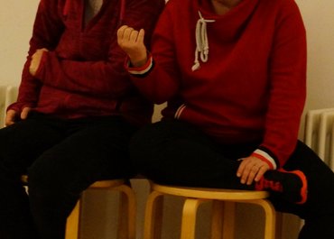 Zwei Frauen sitzen auf einem Hocker, eine lächelt, die andere spricht.