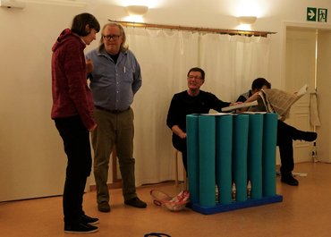 Zwei Personen stehen links nebeneinander und sprechen, rechts sitzen zwei Personen, von denen einer nach links schaut und der andere in die Zeitung liest.