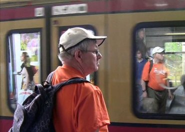 Mann mit Kappe und Rucksack steht mit dem Rücken zur Kamera vor einer einfahrenden Straßenbahn.
