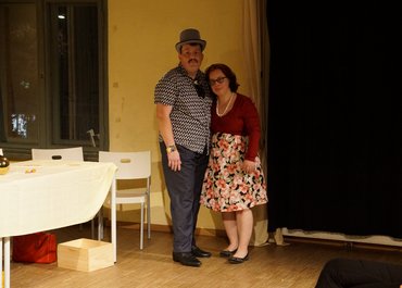 Ein Darsteller mit Hut und eine Darstellerin im Kleid stehen nebeneinander und umarmen sich halbseitig.