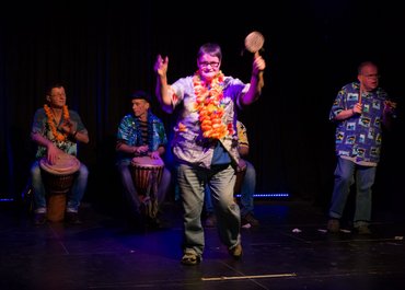 Person auf der Bühne mit Blumenhalskette, im Hintergrund eine Trommelgruppe.