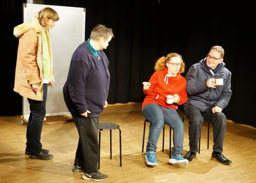 Zwei Darsteller sitzen auf Hockern mit Tassen in der Hand, einer zeigt auf zwei kommende Darsteller.