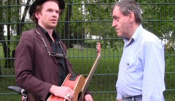 Mann mit Gitarre blickt in die Umgebung, während ein anderer Mann ihn anschaut.