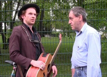 Mann mit Gitarre blickt in die Umgebung, während ein anderer Mann ihn anschaut.