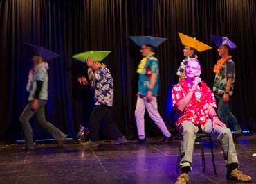 Person sitzt im Vordergrund auf einem Stuhl mit Mikrofon in der Hand, im Hintergrund Personen mit Hütchen auf dem Kopf, die ein Schiff formen.