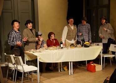 Sieben Personen am Esstisch, fünf stehen singend, zwei sitzen mit gesenktem Kopf und Hand vor dem Gesicht.